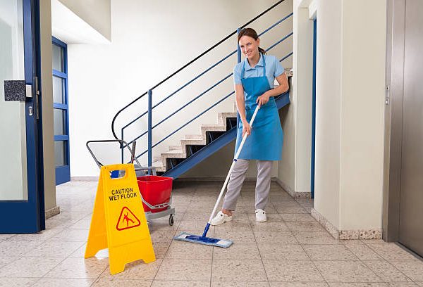 Young Happy Female Worker With Cleaning Equipments And Wet Floor Sign On Floor